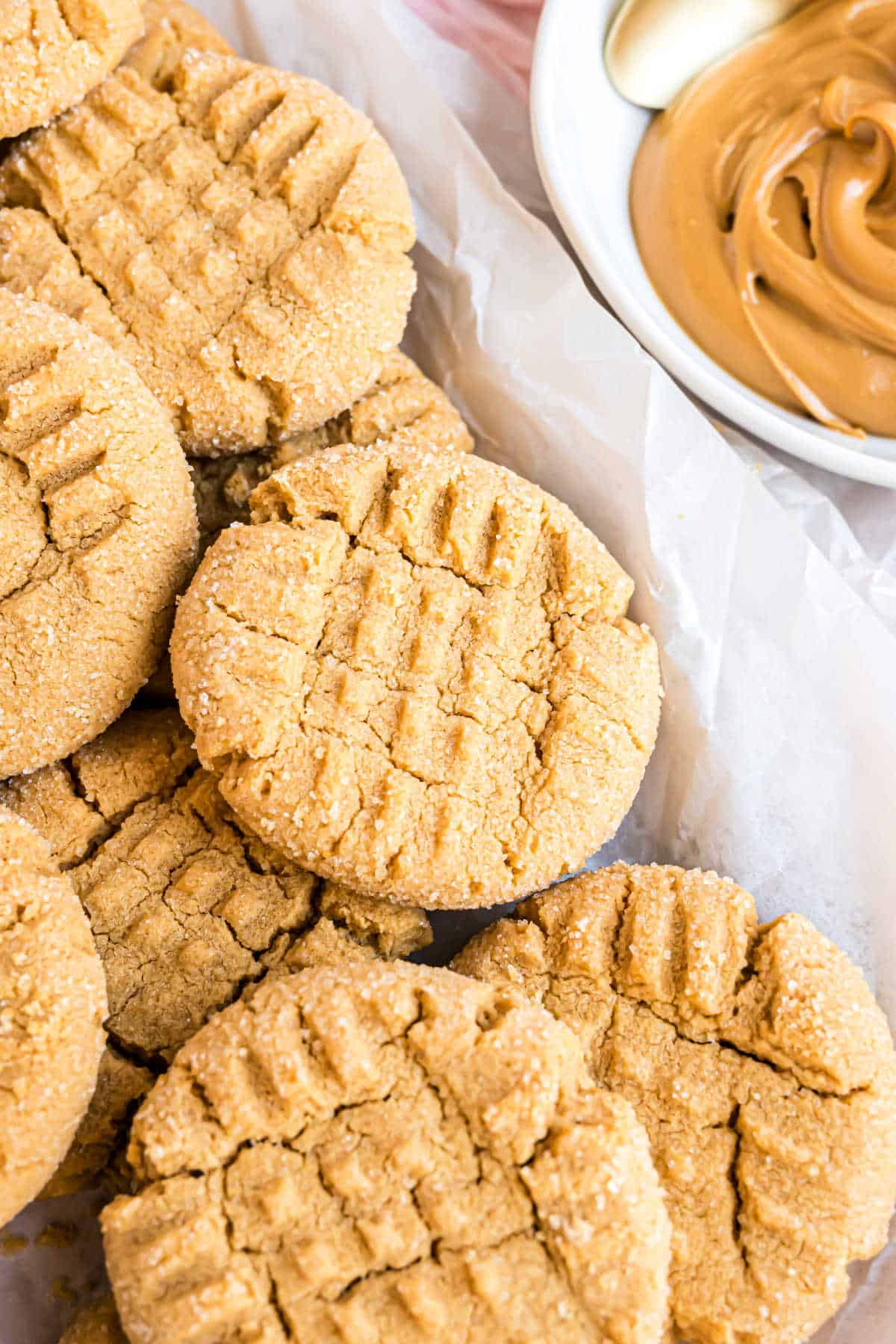 A batch of peanut butter cookies with crisscross tops sits beside a bowl of creamy peanut butter, showcasing the perfect peanut butter cookies recipe.