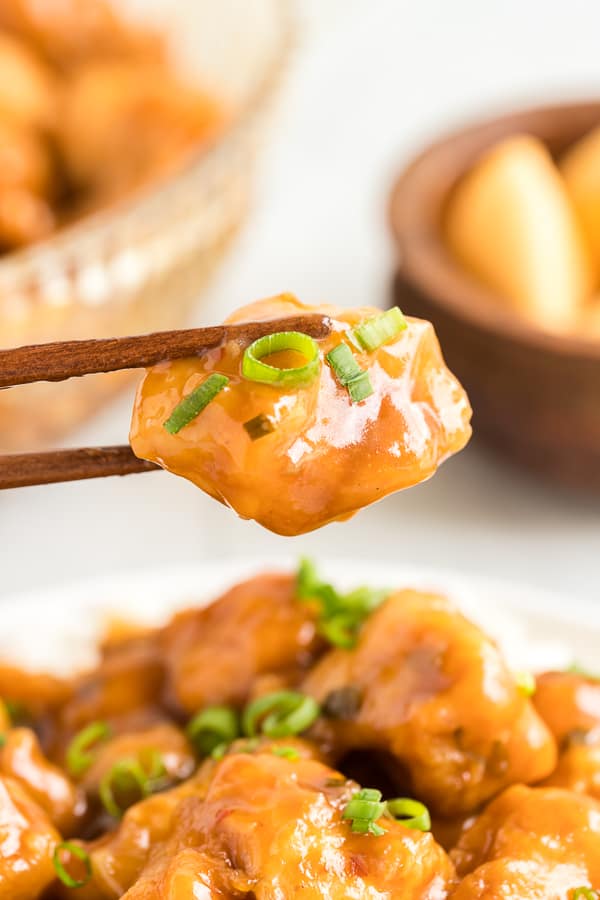 Piece of glazed orange chicken with green onions being held by chopsticks.