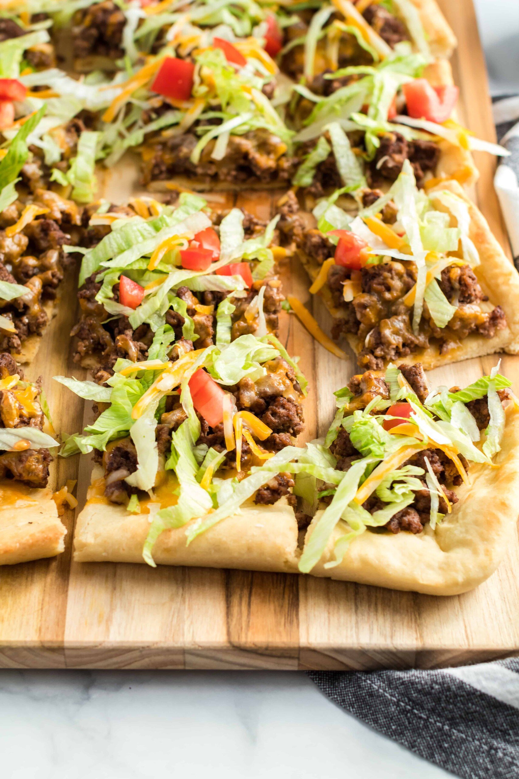 Beef taco pizza with lettuce and tomato cut on a wooden cutting board.