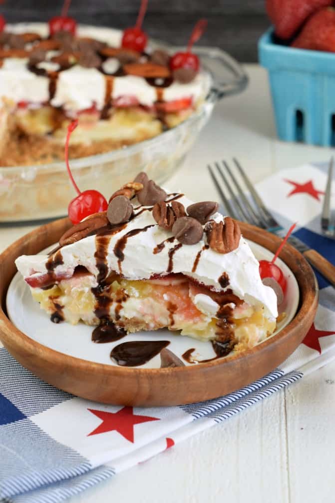 Banana Split cheesecake with layers of graham cracker crust, pineapple, strawberries, and banana with whipped cream, chocolate chips, pecans, and a cherry.