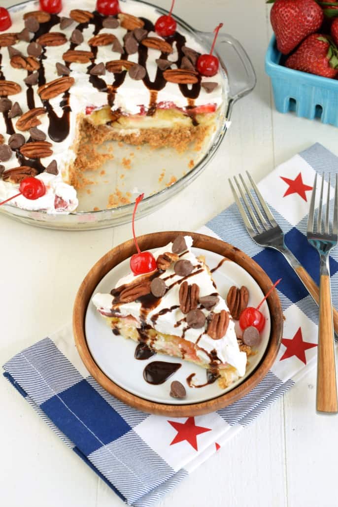 Slice of cheesecake topped with whipped cream, chocolate, pecans, and cherry on a white plate with red, white, and blue napkin.