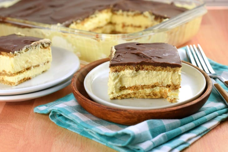 This Eclair Cake recipe is an old fashioned icebox cake with layers of graham crackers, pudding, and chocolate frosting. An easy no bake dessert recipe! Slice of chocolate frosted icebox cake on a white plate.