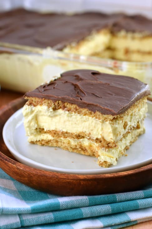 One square slice of chocolate eclair cake with layers of graham cracker, pudding, and chocolate frosting, on a white plate and teal napkin.