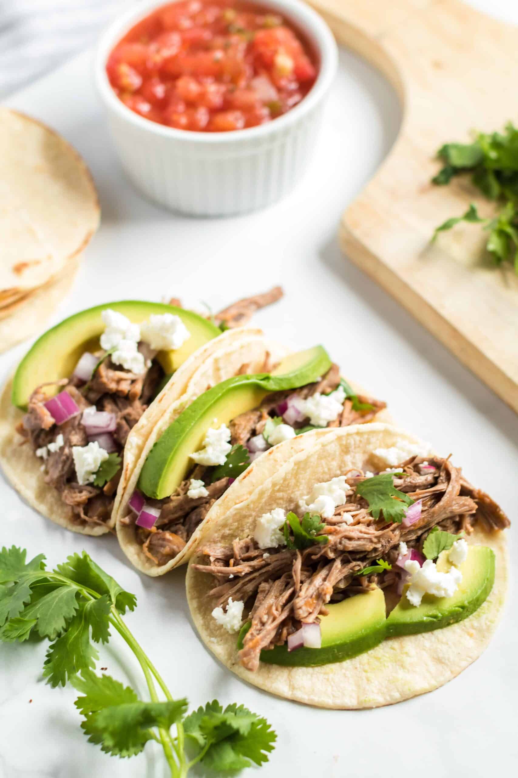 Slow Cooker Barbacoa Beef on flour tortilla with cilantro, cheese, and avocado with a side of fresh salsa.