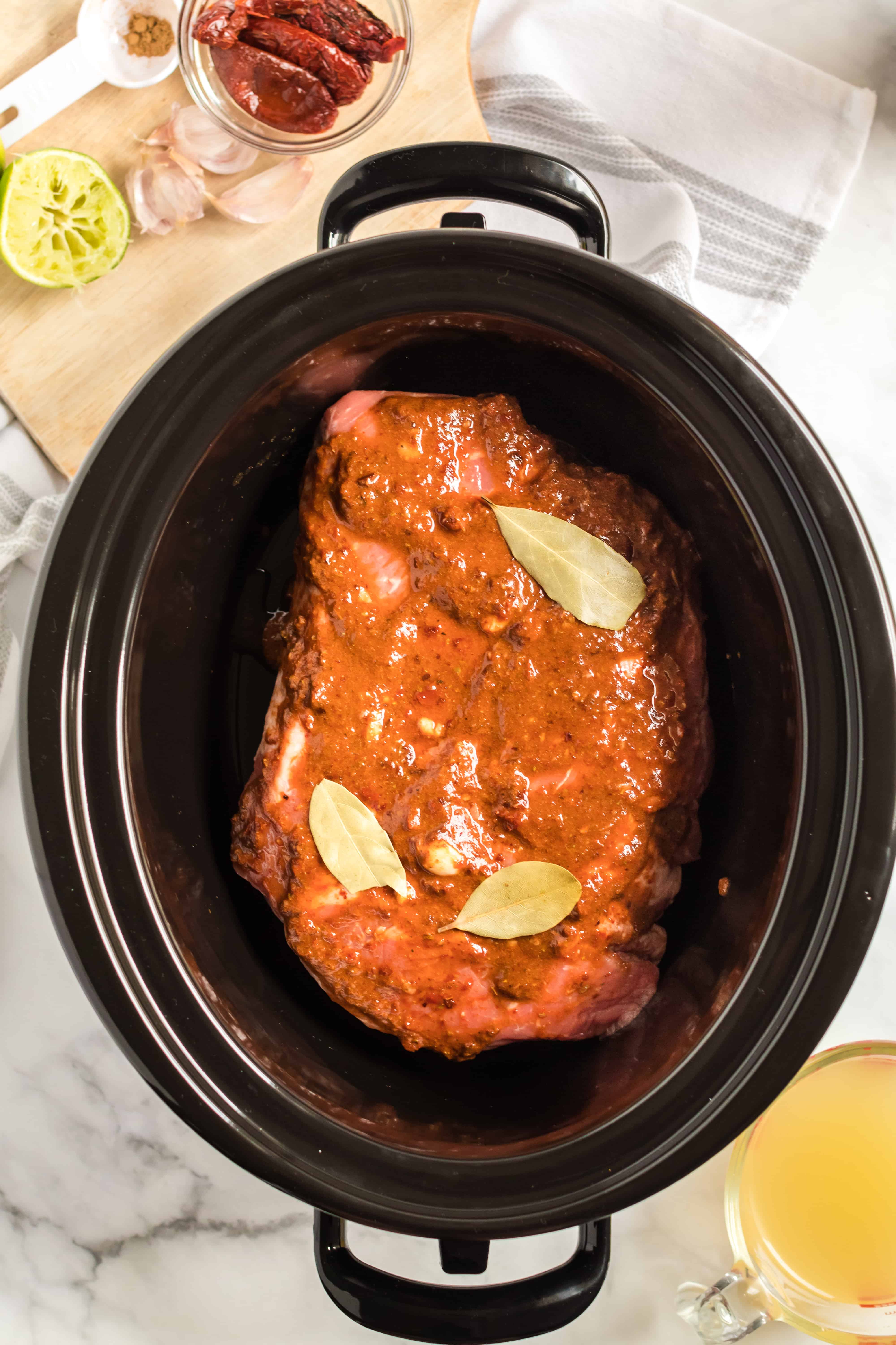 Crockpot with large cut of chuck steak topped with barbacoa sauce and bay leaves.