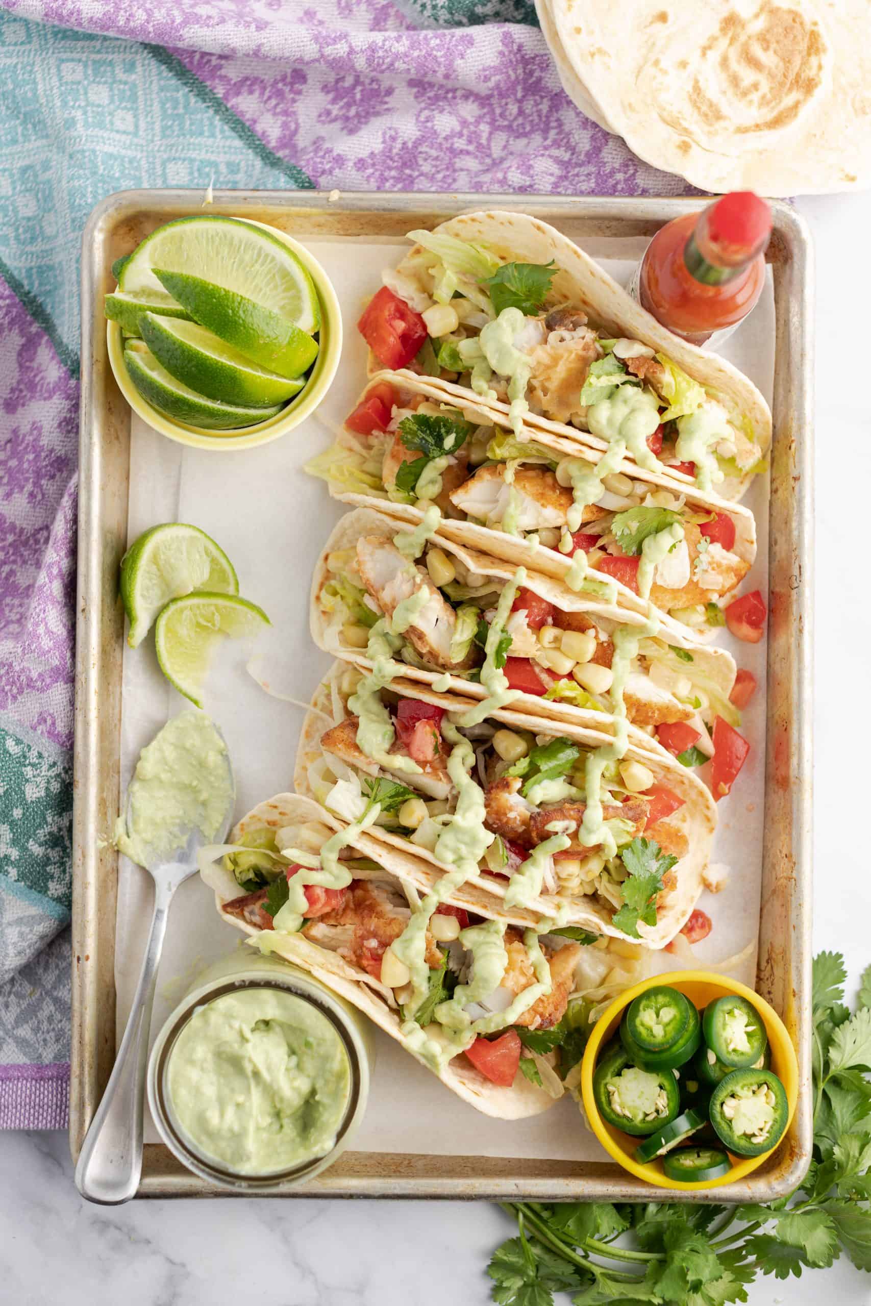 Metal baking sheet with fish tacos on flour tortillas drizzled with avocado sauce.