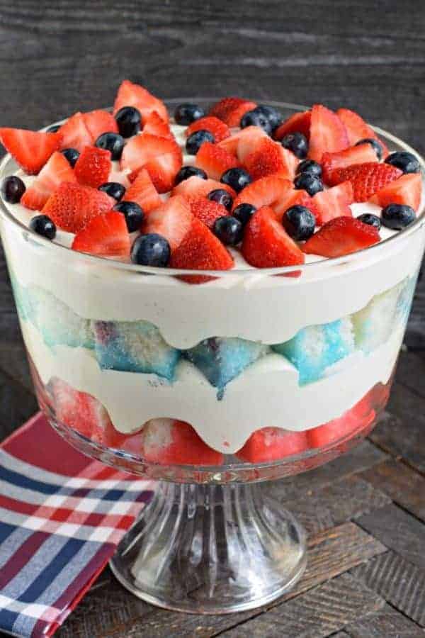 Clear glass trifle bowl filled with layers of blue jello cake, red jello cake, and white cheesecake filling. Topped with fresh berries.