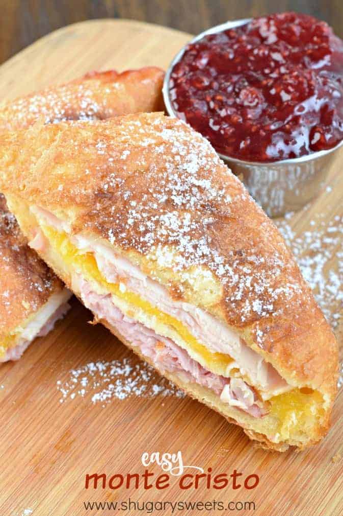 Wooden cutting board with half a slice of monte cristo sandwich (turkey, cheese, ham). Deep fried and sprinkled with powdered sugar and a side of raspberry jam.