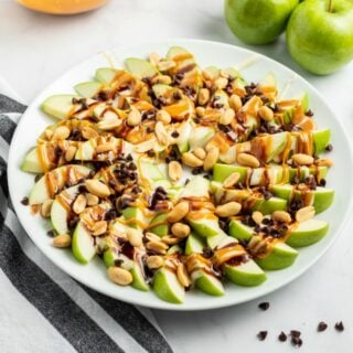 Green apple slices arranged on a white plate in a spiral and topped with melted marshmallow, chocolate, caramel, and peanuts.