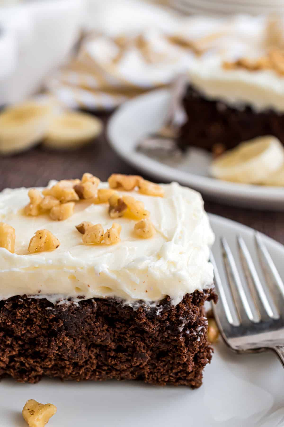 Slice of chocolate banana cake topped with cream cheese frosting and walnuts.