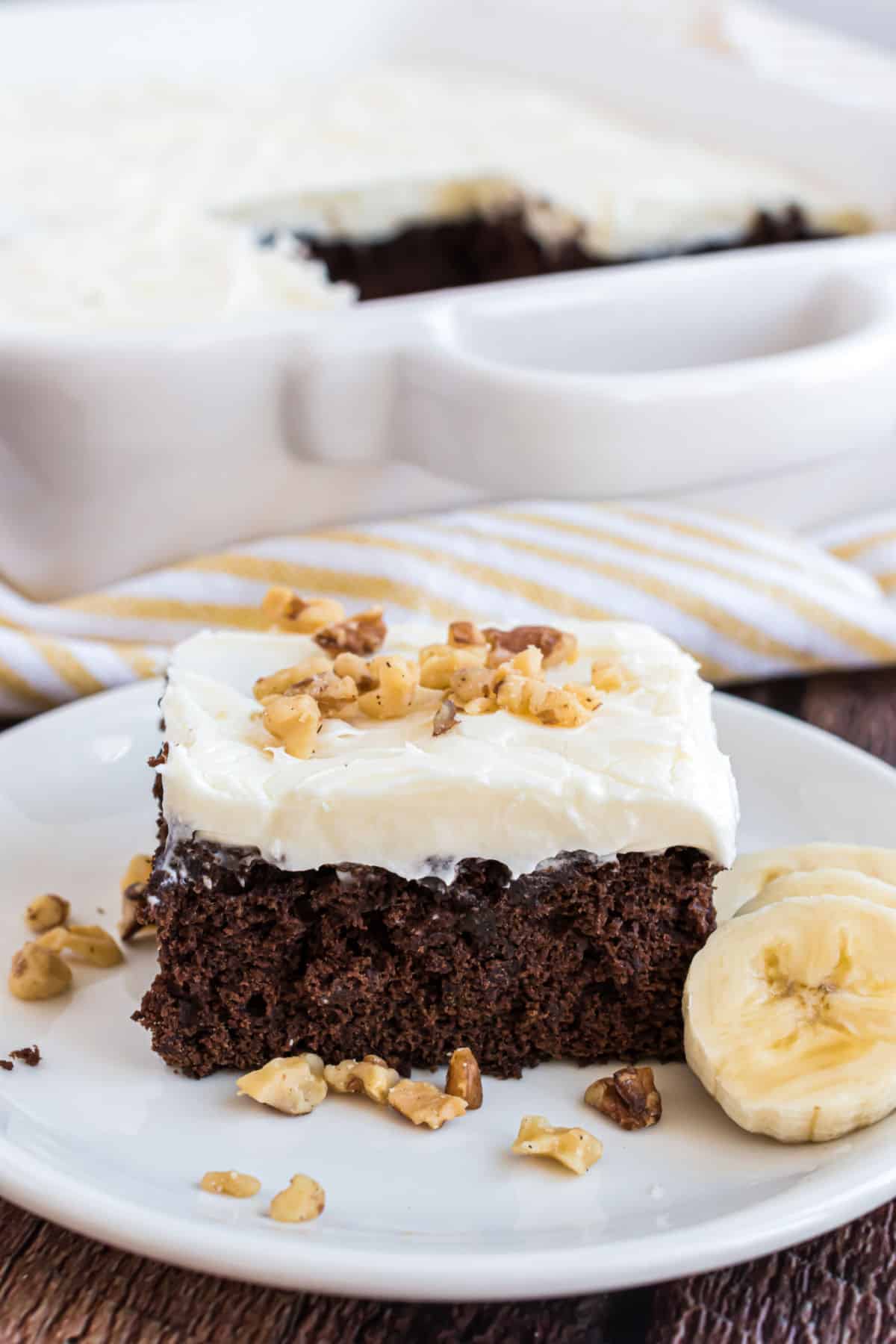 Chocolate banana cake with cream cheese frosting and walnuts served on a white plate.