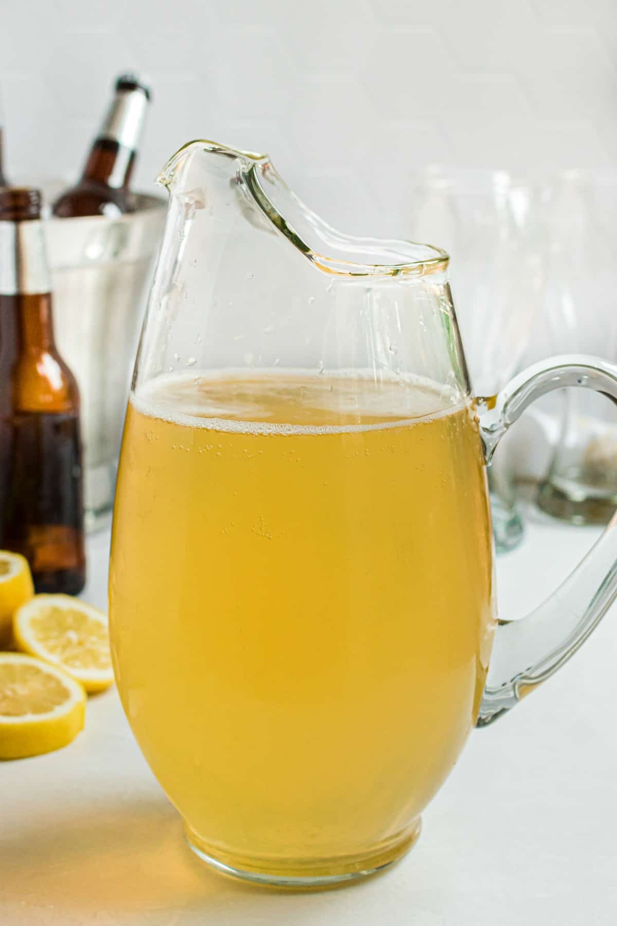 Clear glass pitcher of homemade lemon shandy with bottles of beer in background.