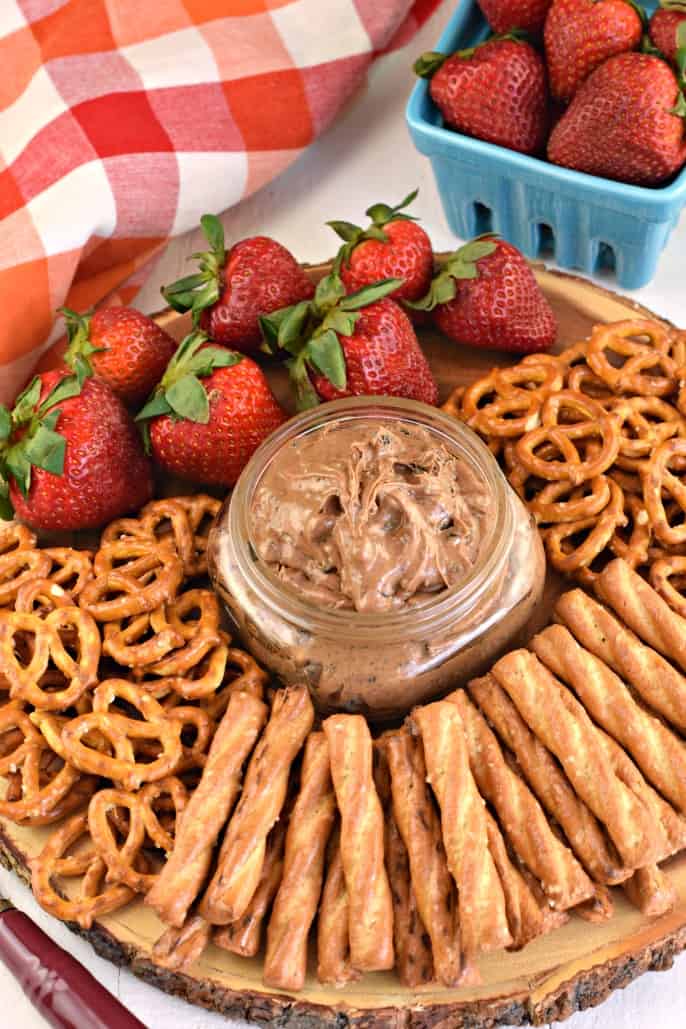 Small clear glass mason jar with oreo peanut butter dip on a wooden serving plate with strawberries and pretzels.