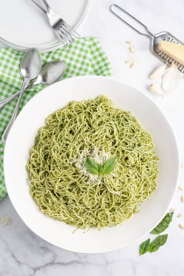 White bowl with basil pesto pasta, fresh grated parmesan cheese, and a side of plates and forks.