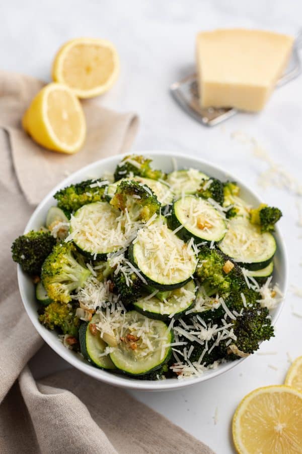 White bowl of roasted broccoli and zucchini topped with parmesan cheese and fresh lemon juice