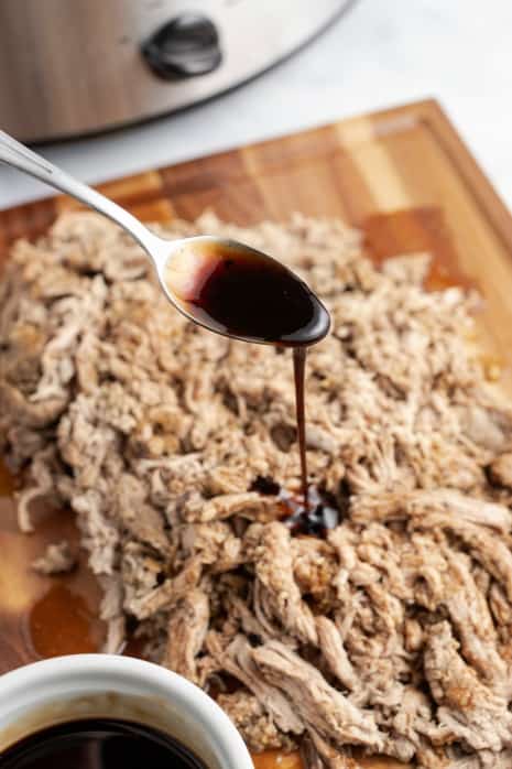 Wooden cutting board with shredded pork tenderloin. Metal spoon drizzle balsamic glaze over the top with crockpot in background.