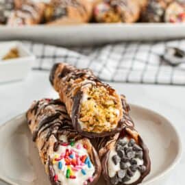 Three cannolis on a white dessert plate, each dipped in either pistachios, chocolate chips, or sprinkles.