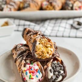 Three cannolis on a white dessert plate, each dipped in either pistachios, chocolate chips, or sprinkles.