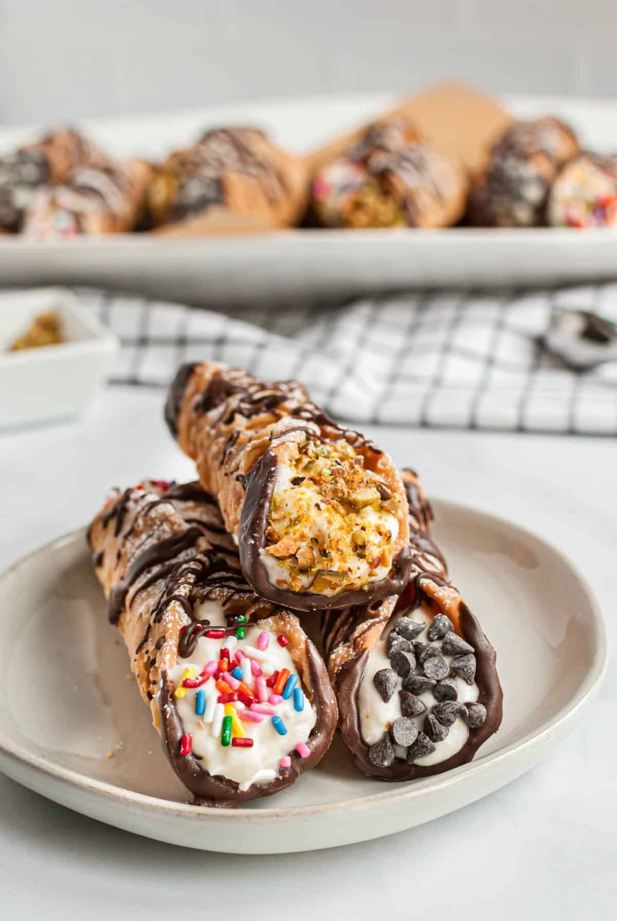 Three cannolis on a white dessert plate, each dipped in either pistachios, chocolate chips, or sprinkles.