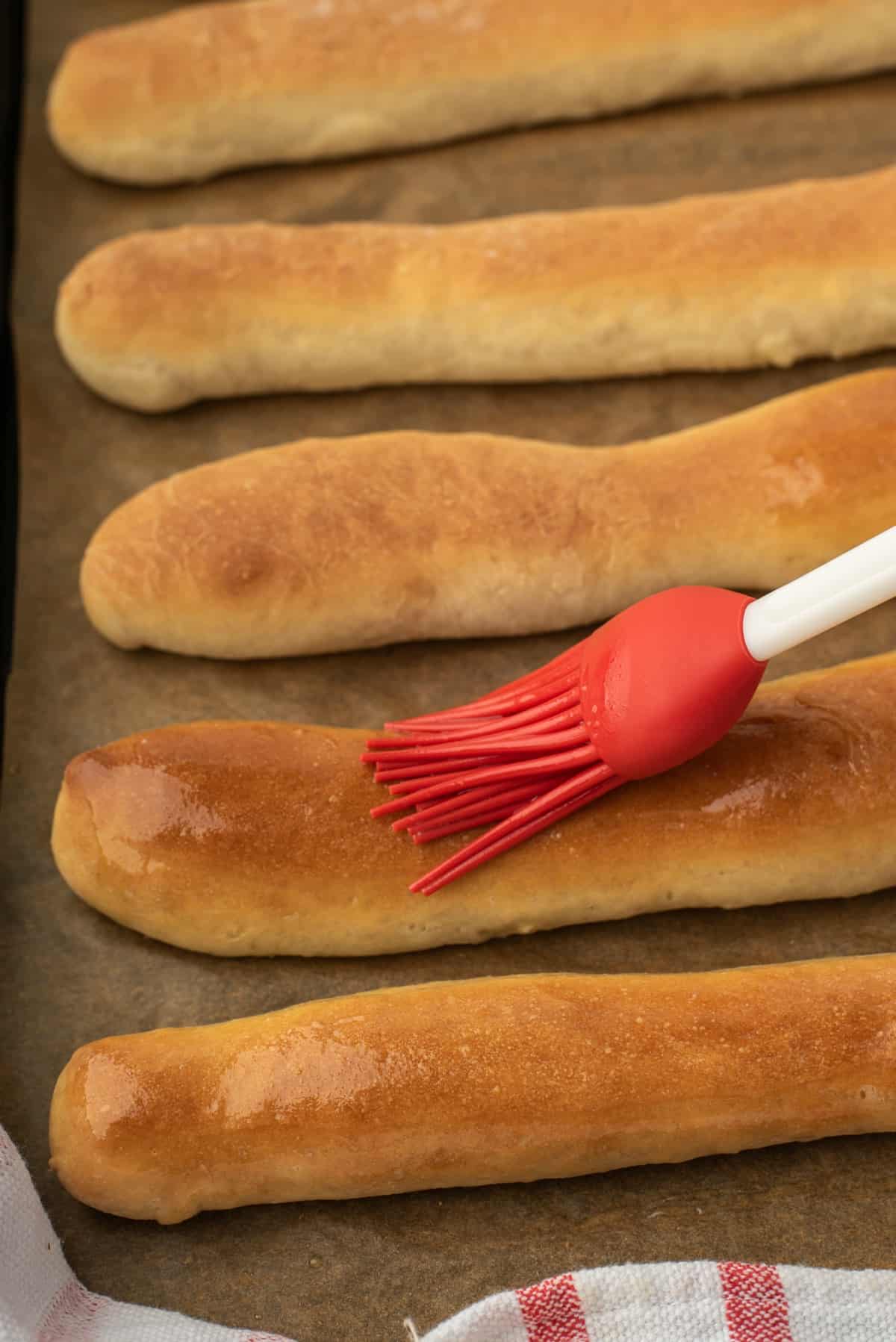 Baked breadsticks on a metal baking sheet being brushed with butter using a red pastry brush.