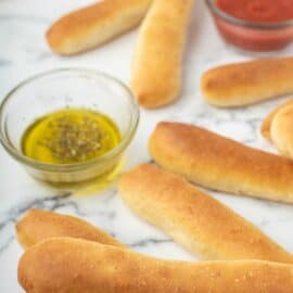 Marble counter with breadsticks laying on top with small bowls of marinara and oil.