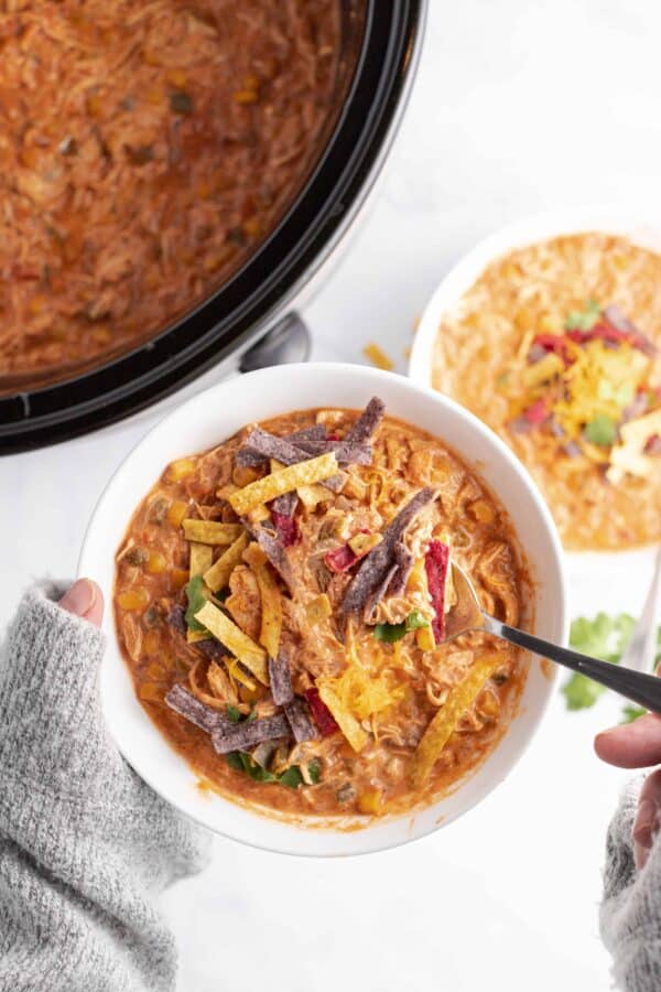 Warming and hearty Chicken Taco Soup cooks up easily in the slow cooker. Topped with crispy tortilla strips and shredded cheese, this is an easy meal the whole family will love. White bowl with chicken taco soup topped with cheese and colored tortilla strips