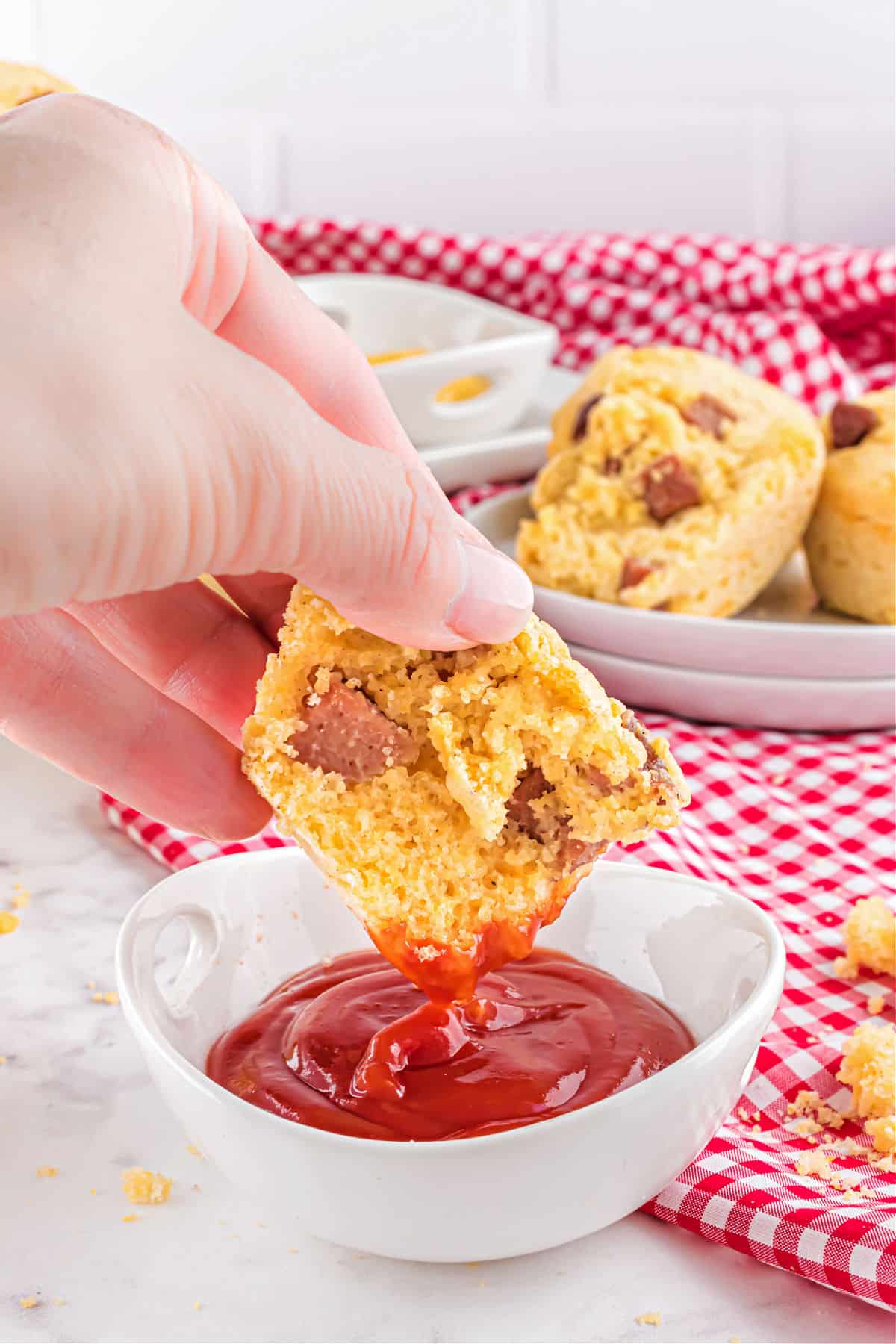 Half a corn dog muffin being dipped into a white bowl with ketchup.