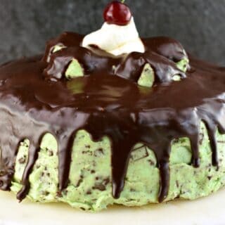 Chocolate cake with mint chip frosting and chocolate ganache. On a white cake plate and topped with a cherry.