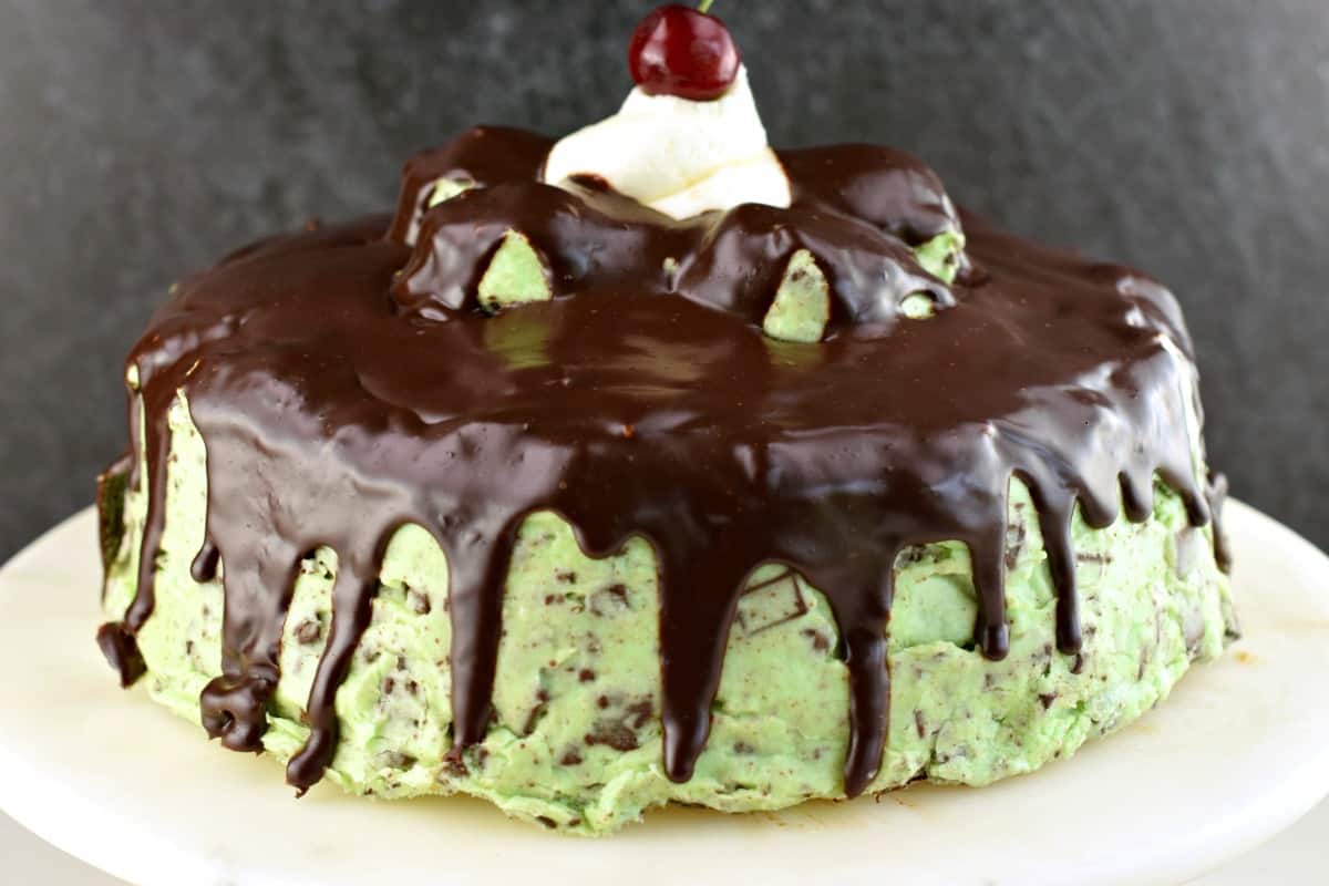 Chocolate cake with mint chip frosting and chocolate ganache. On a white cake plate and topped with a cherry.