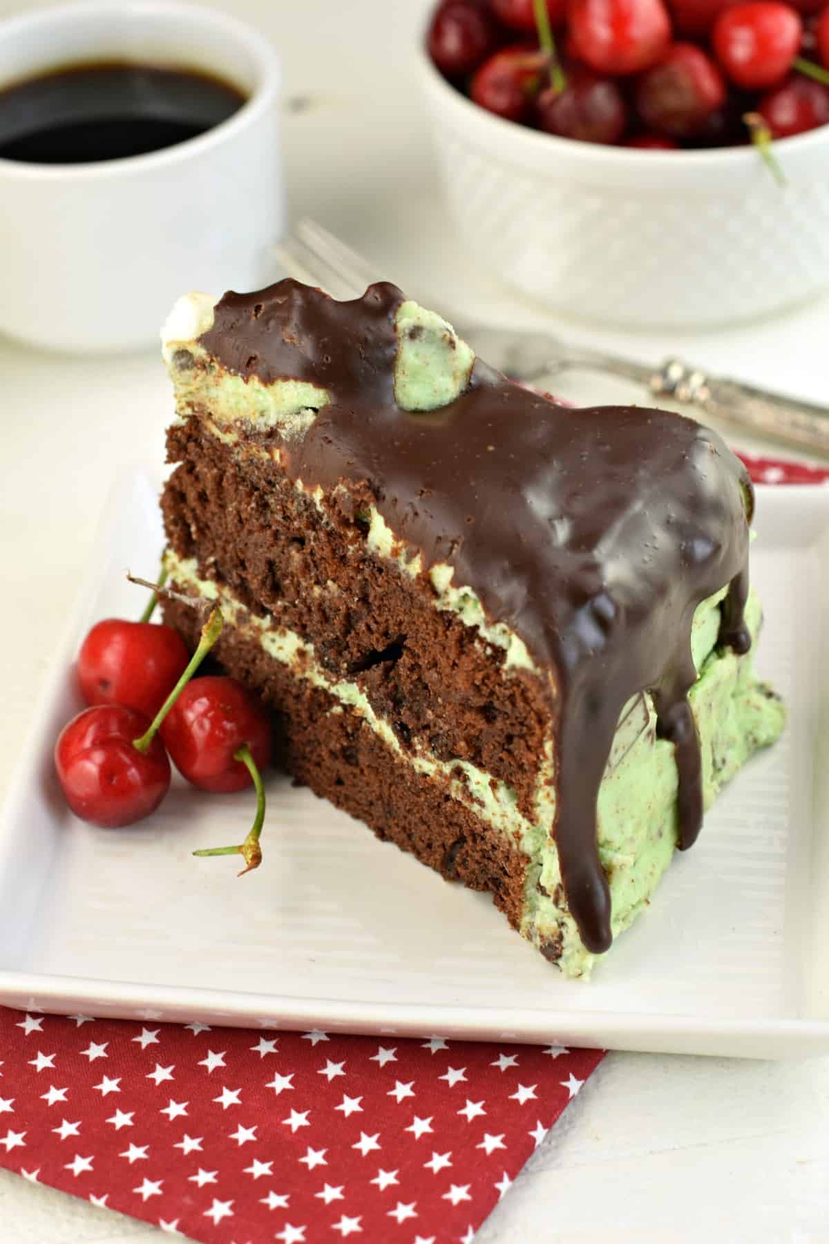 Thick slice of chocolate layer cake with mint chocolate chip frosting tinted green and covered in chocolate ganache. Served on a white square dessert plate with fresh cherries.