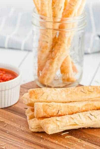 Puff pastry bread sticks on wooden bread board with a jar of extra breadsticks in background.