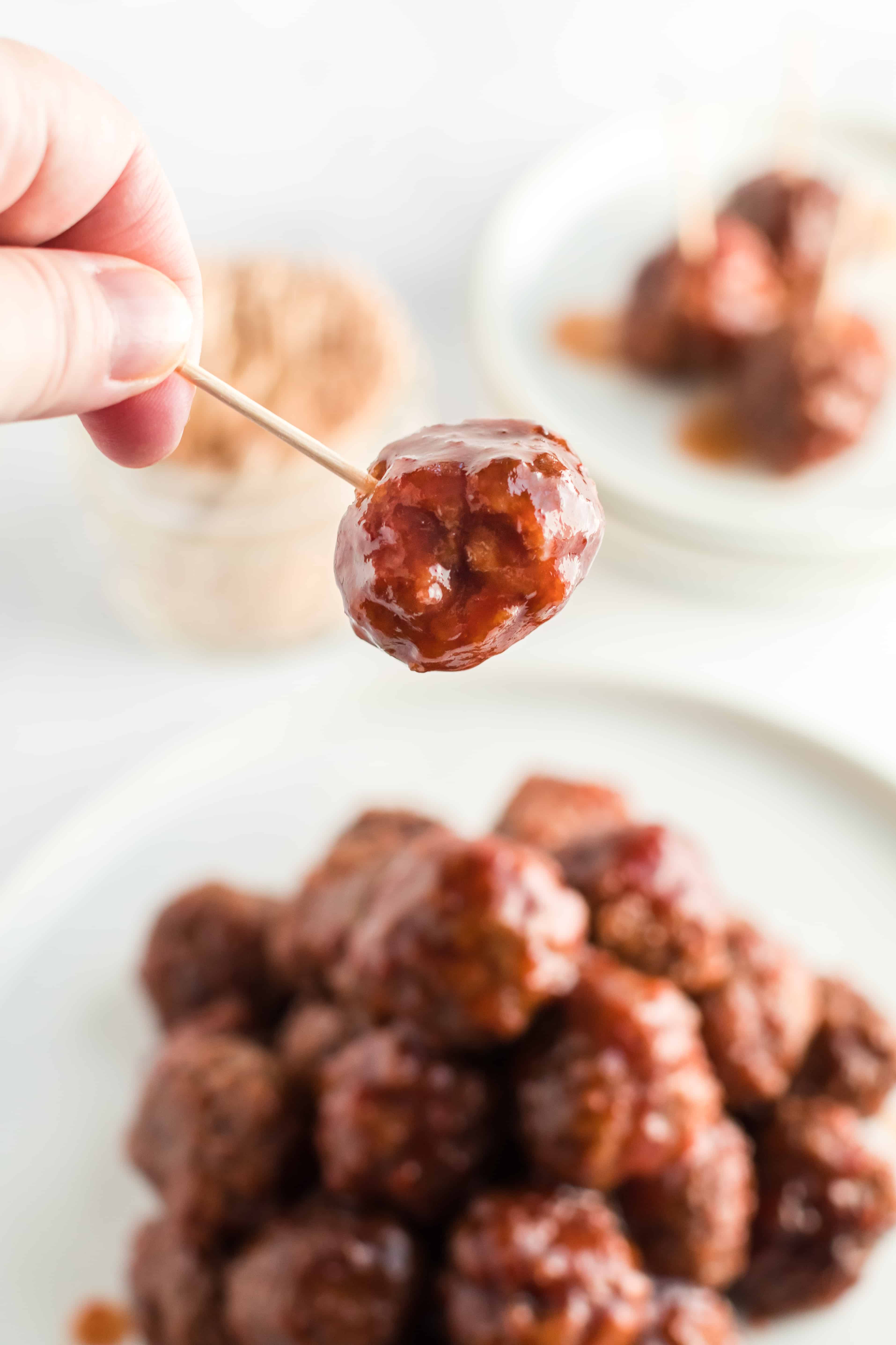 Closeup of toothpick with grape jelly meatballs. Plate of meatballs in background,