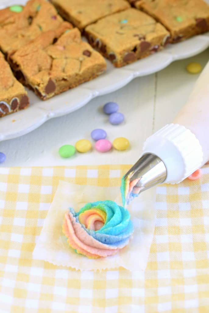 Rose swirl of rainbow frosting on a square of parchment paper with frosting bag and tip next to it. M&M's in background with cookie bars.