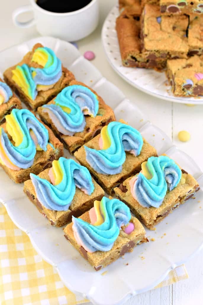 Plate of square cute rainbow frosted chocolate chip cookies bars with a plate of plain (non frosted) bars in the back ground.