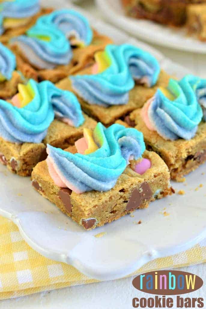 Squares of chocolate chip cookie bars on a white plate. Frosted with a swirl of rainbow frosting.