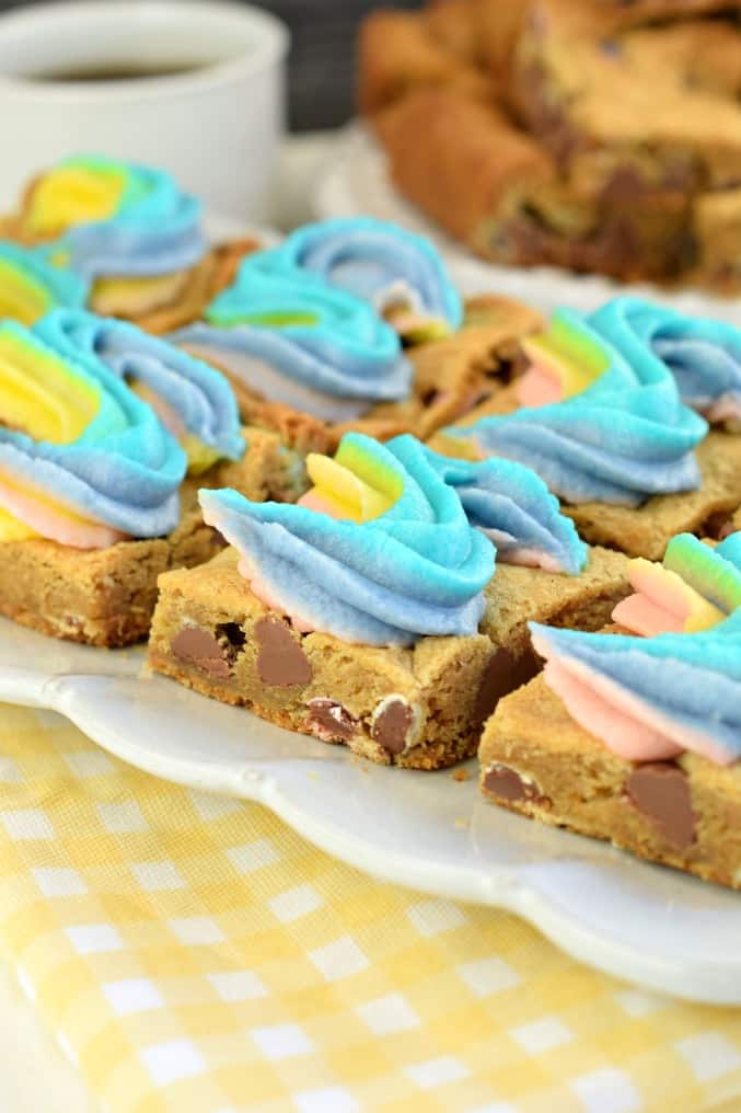 Chocolate Chip cookie bars with rainbow swirled frosting on a white plate with yellow napkin underneath.