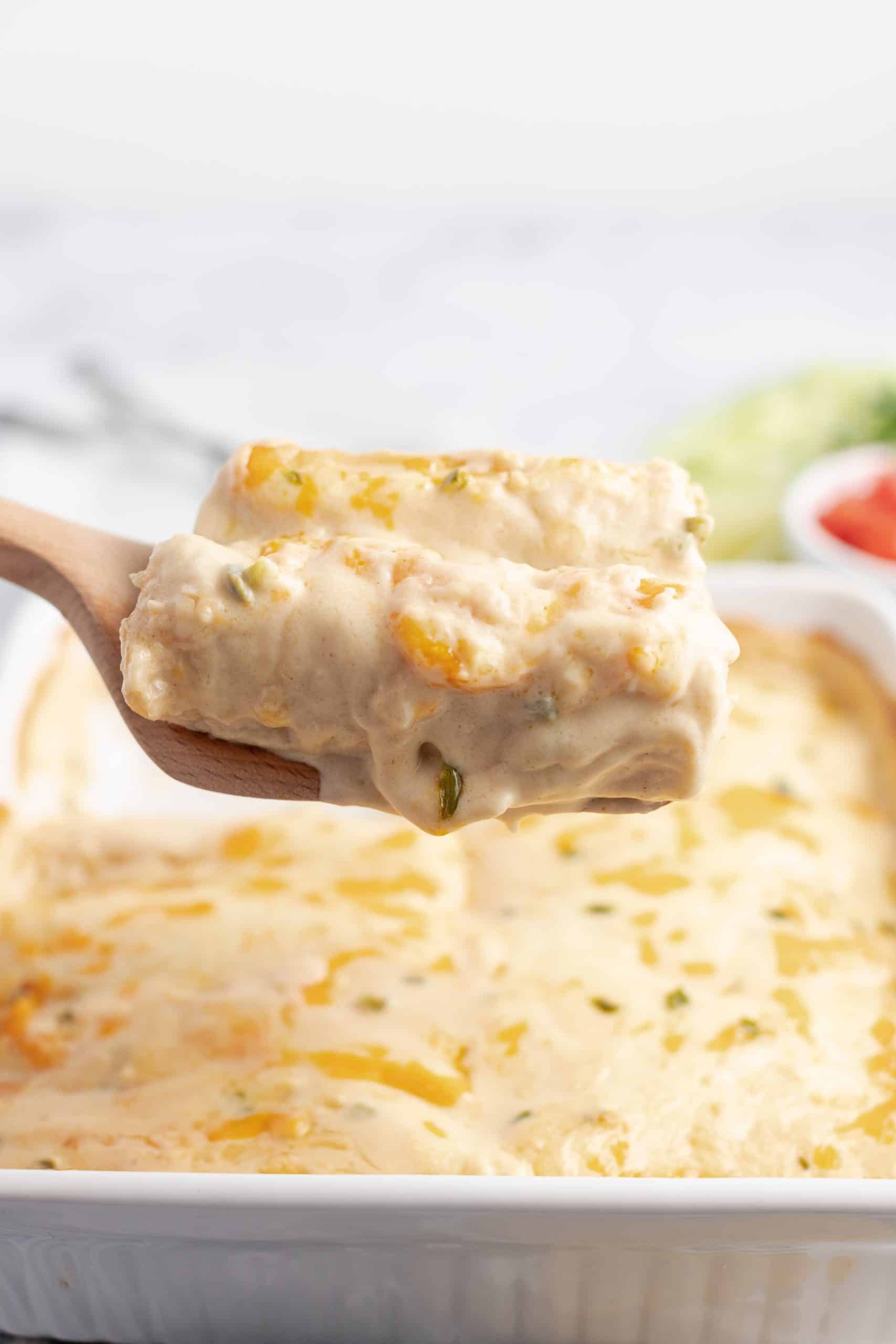 Two creamy enchiladas being lifted out of a 13x9 baking dish with a wooden spatula.