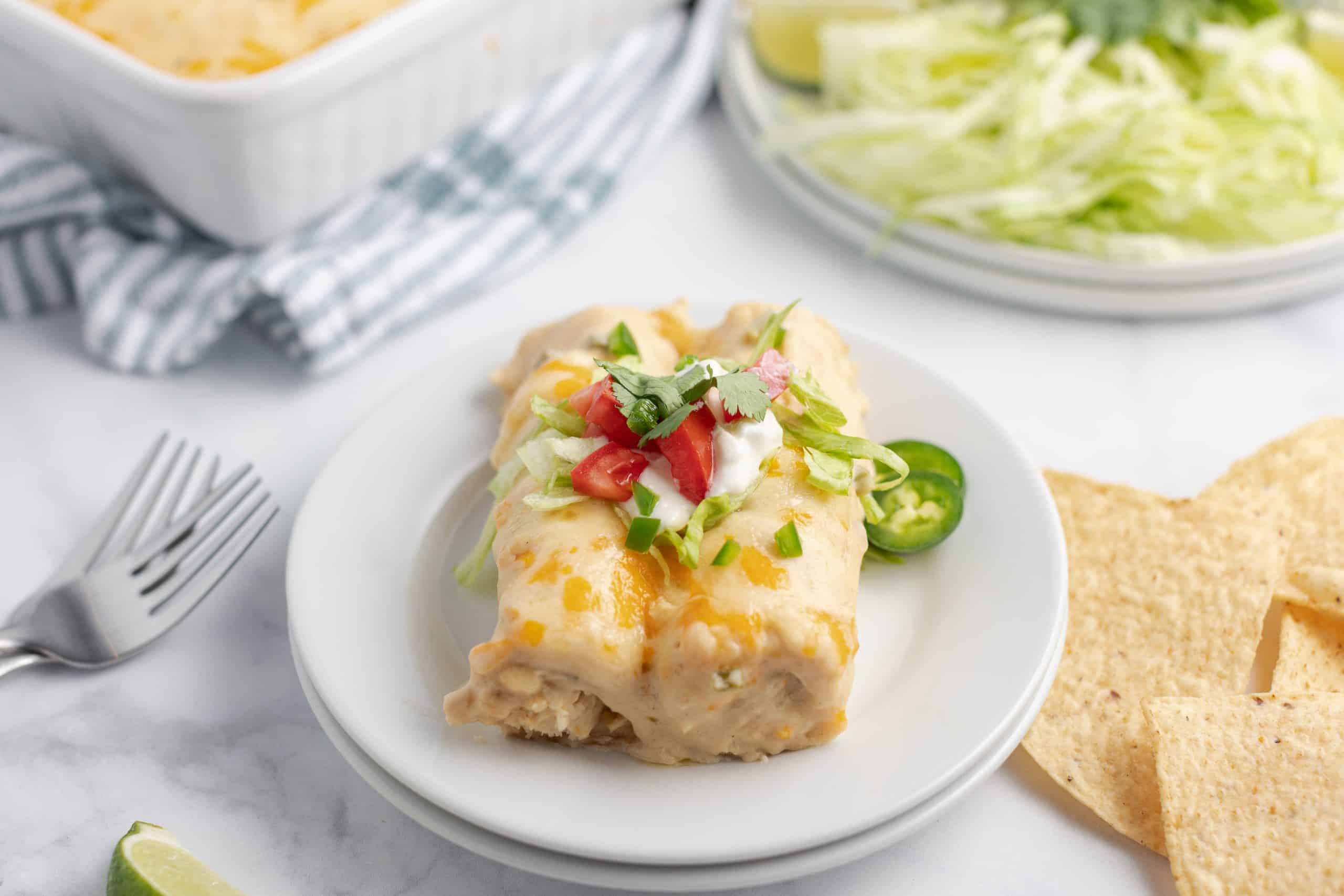 White plate with two enchiladas topped with sour cream, tomato, jalapeno and cilantro.