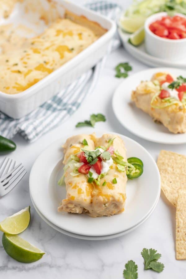 Two creamy lime chicken enchiladas on a white plate with jalapeno, cilantro, tomato, lime wedges, and sour cream. Pan of enchiladas and topping in background