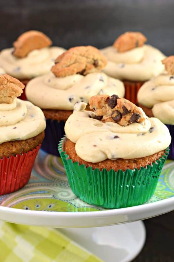 Multiple Chocolate chip cupcakes with cookie dough frosting. Topped with chocolate chip cookie on a cake plate.