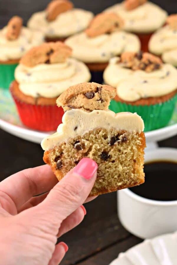 Half of a chocolate chip cupcake in a hand. Topped with cookie dough frosting and a chocolate chip cookie