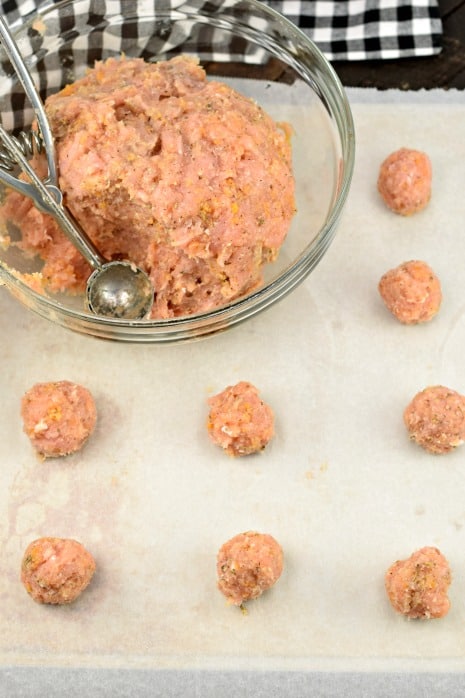 Clear glass bowl with meatball mixture being scooped onto parchment paper with a metal scoop.