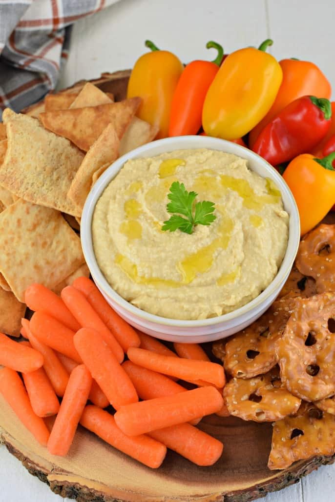 Hummus with olive oil in a white bowl and served with veggies, pretzels, and pita chips.