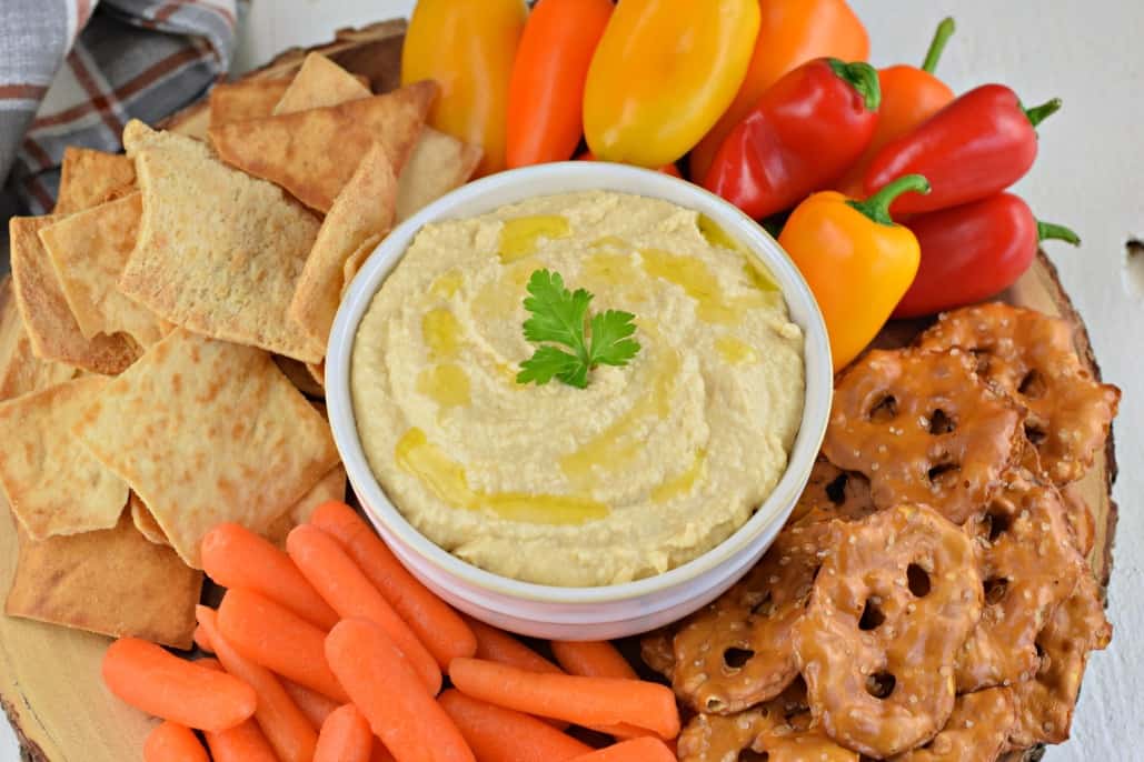 Hummus in a bowl and served on a platter with pretzels, pita chips, and vegetables.