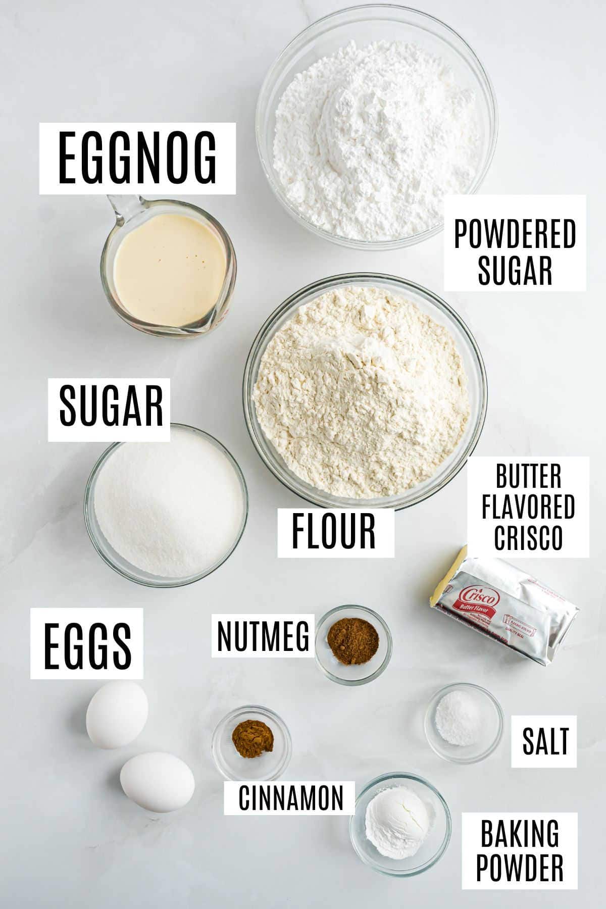 Baking ingredients for eggnog muffins, including eggs, flour, sugar, spices, and Crisco, are labeled and arranged on a white surface.