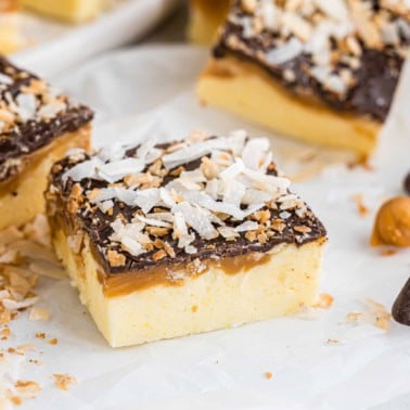 A Samoa fudge dessert bar with a yellow base, caramel, a rich chocolate layer, and coconut flakes on top.