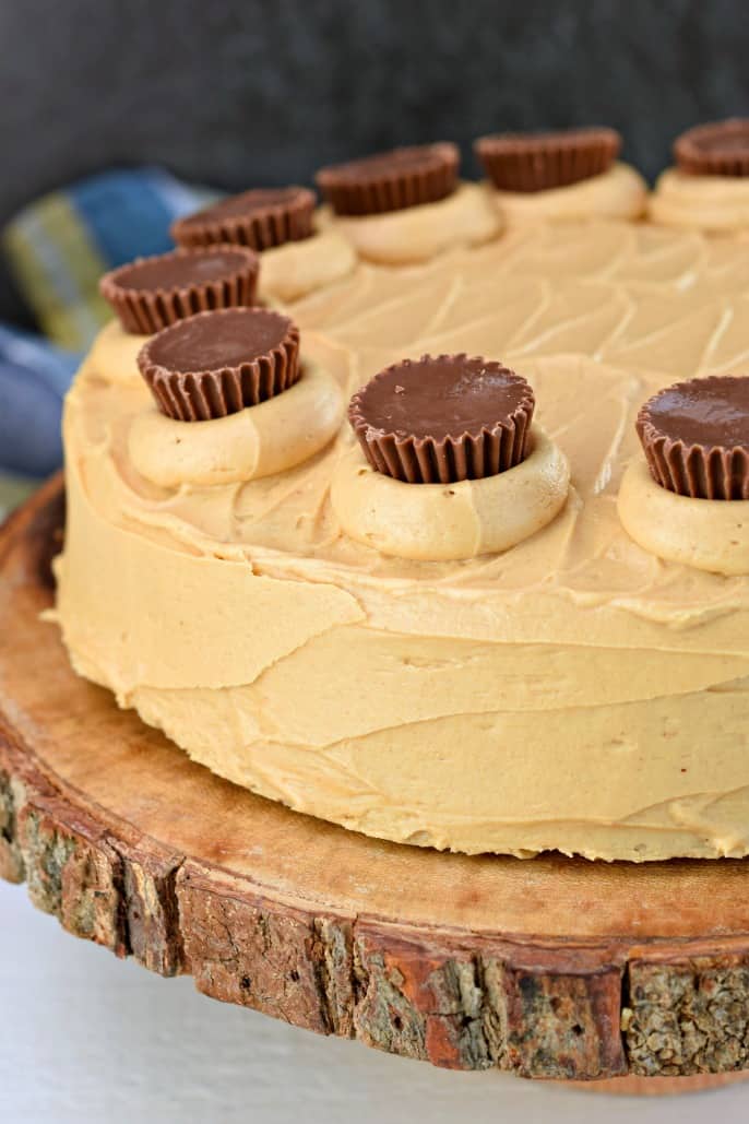Peanut butter frosted cake on a wooden cake plate and garnished with mini Reese's peanut butter cups.