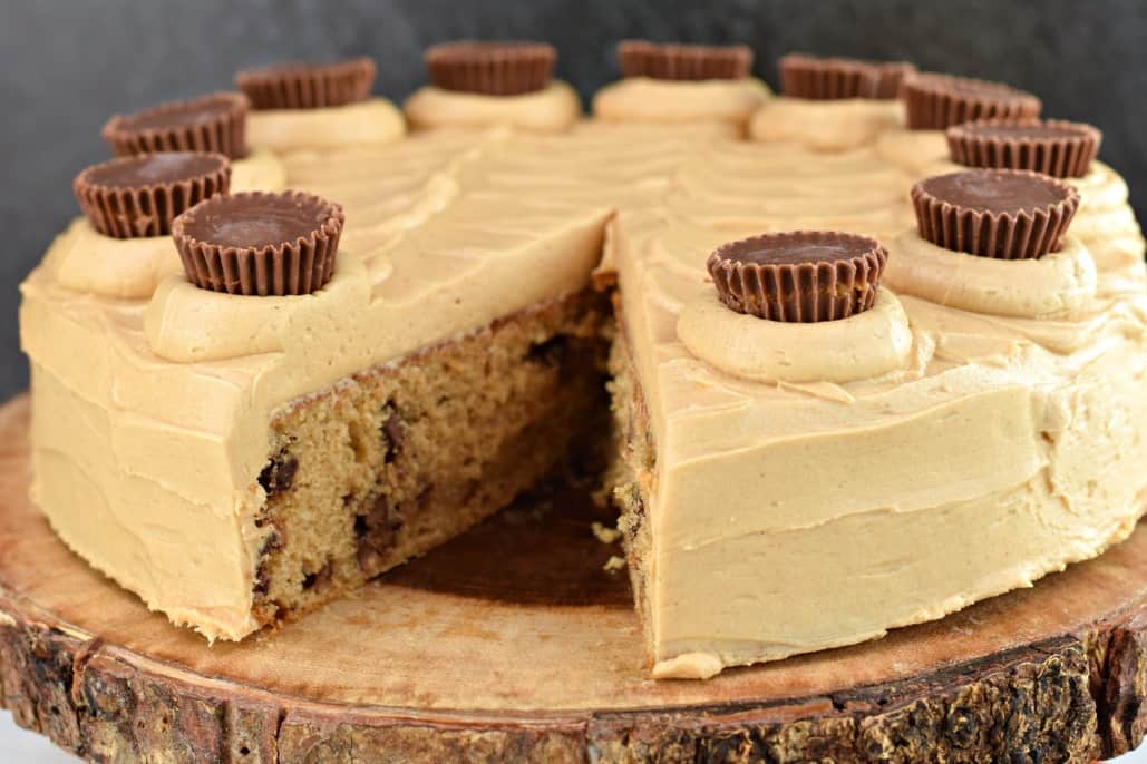 Peanut butter cake with frosting and Reese's peanut butter cups on top, served on a wooden cake platter.