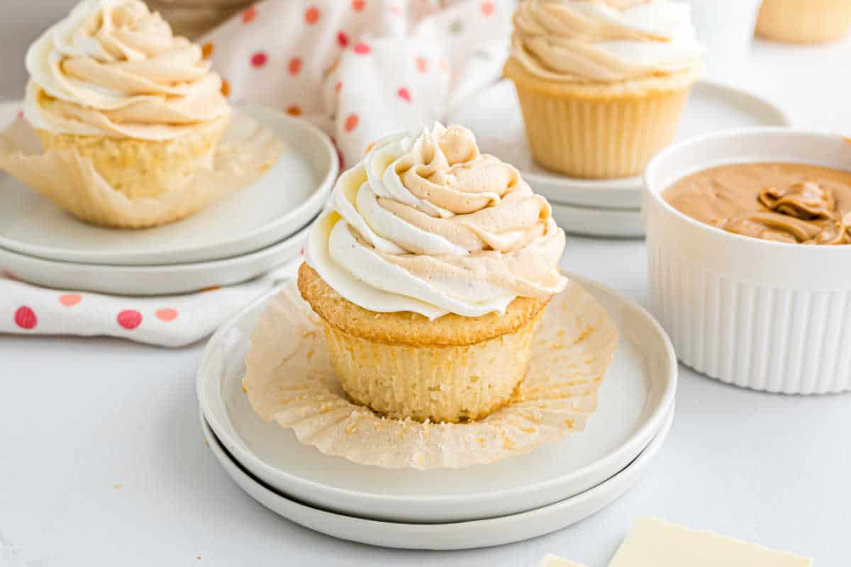Vanilla cupcake with swirled frosting on a plate, surrounded by white chocolate cupcakes and a bowl of peanut butter nearby.