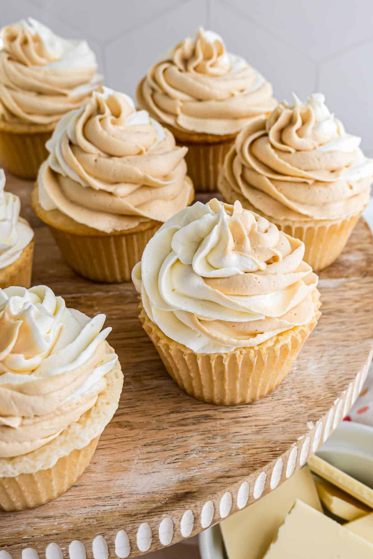 White chocolate cupcakes with vanilla bases, topped with swirled white and light brown frosting, displayed elegantly on a wooden cake stand.